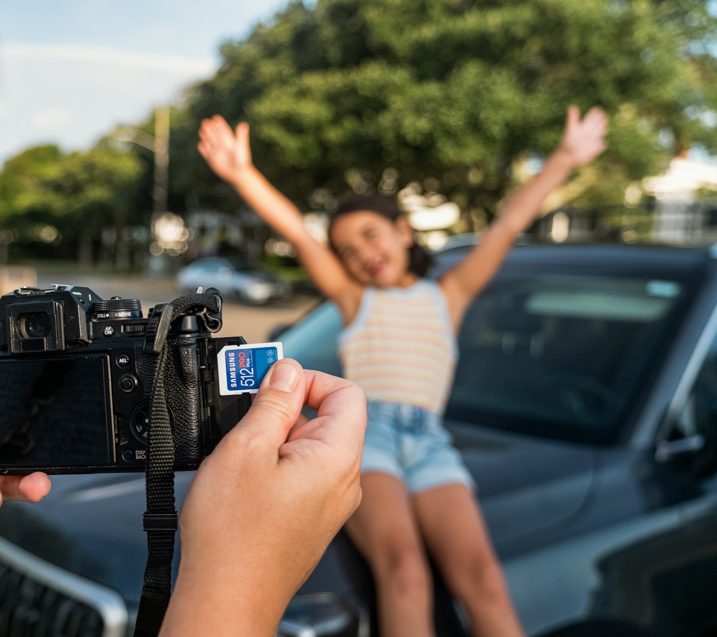 Kuva muistikortin vaihdosta kameraan. Taustalla iloinen lapsi istuu auton päällä ja poseeraa.